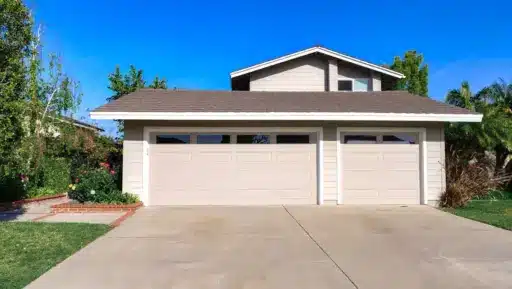 Beige two-car garage with brown shingled roof in residential neighborhood under clear blue sky