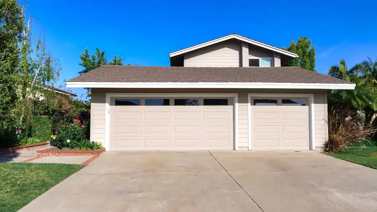 Beige two-car garage with brown shingled roof in residential neighborhood under clear blue sky