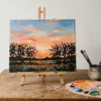 acrylic landscape painting on canvas with sunset sky, flowers, brushes, palette, and paint tubes on wooden table