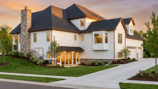 Large modern house with white siding and stone chimney at sunset, surrounded by landscaped yard
