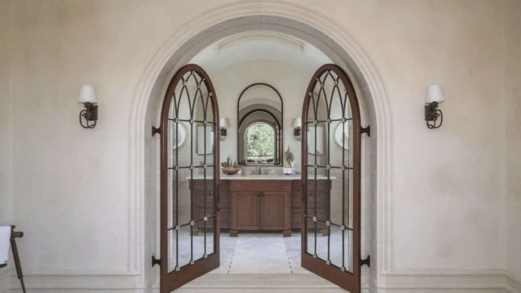 arched double glass doors open into a bathroom with wooden vanity, mirror, and wall sconces framed by a curved entryway