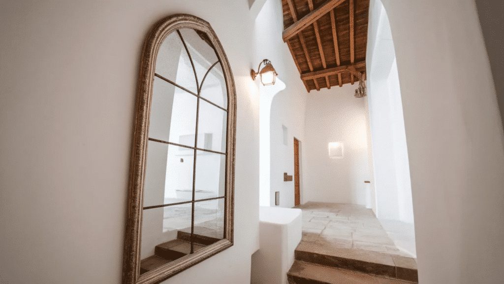 arched hallway with white stucco walls, wood ceiling beams, and tall framed mirror