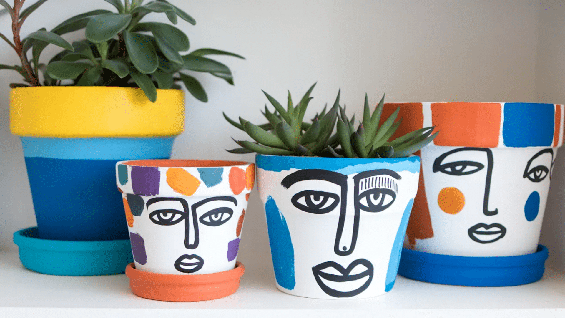 colorful hand-painted plant pots featuring abstract faces and geometric patterns in bright hues like yellow, blue, orange, and purple, with succulents placed inside.