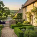 Man in suit presenting charming stone cottage with rose-covered facade and lush green garden