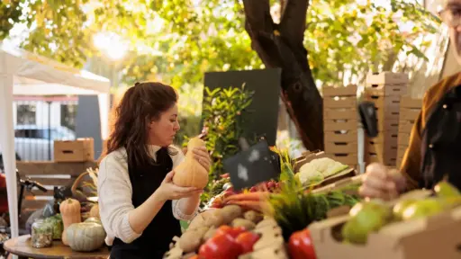 Woman examining butternut squash at an outdoor market stall with fresh produce