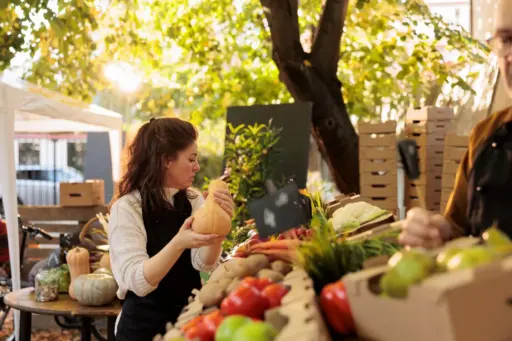 Woman examining butternut squash at an outdoor market stall with fresh produce
