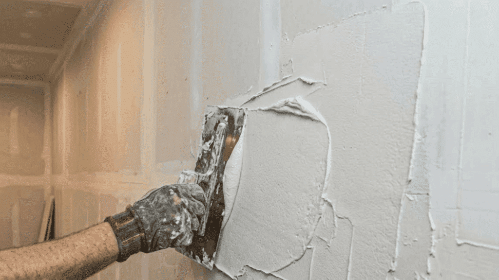 gloved hands spreading fresh first coat plaster across a large indoor wall surface using a trowel and loaded hawk board