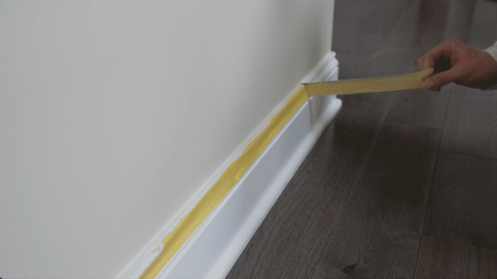 hand placing painter tape along baseboard on wall above wooden floor for clean paint lines