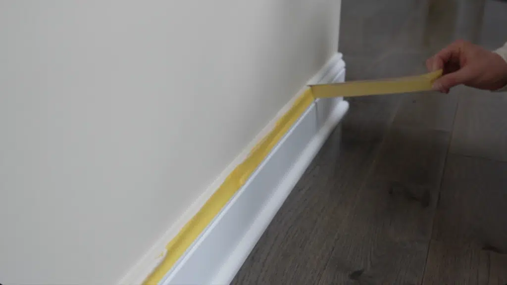 hand placing painter tape along baseboard on wall above wooden floor for clean paint lines