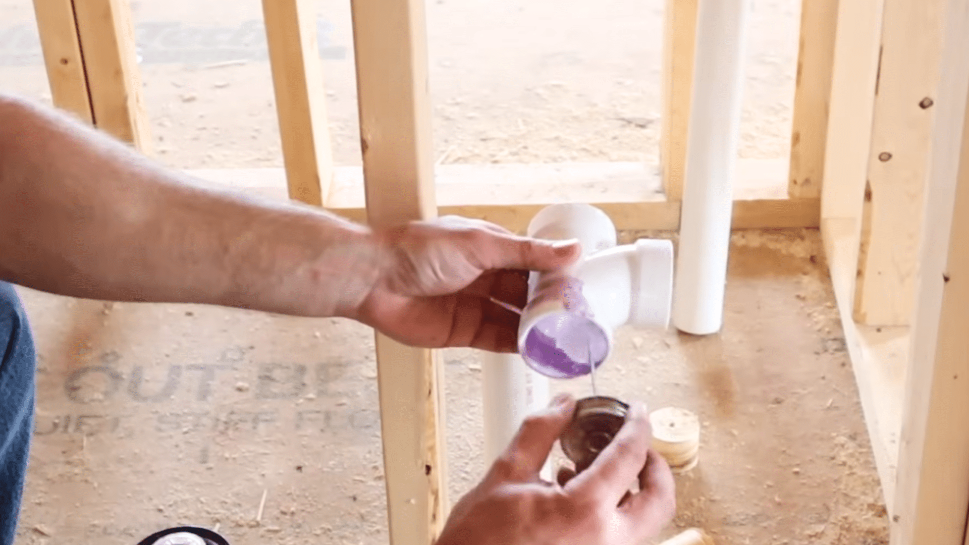 hands applying purple primer to pvc pipe fitting before connecting plumbing pieces inside framed wall structure