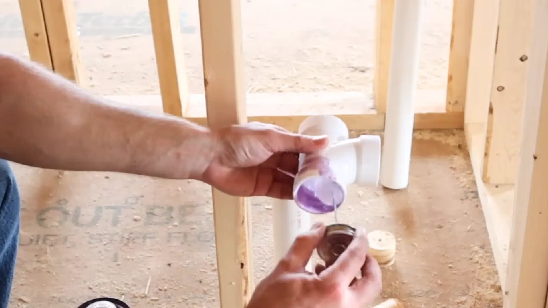 hands applying purple primer to pvc pipe fitting before connecting plumbing pieces inside framed wall structure