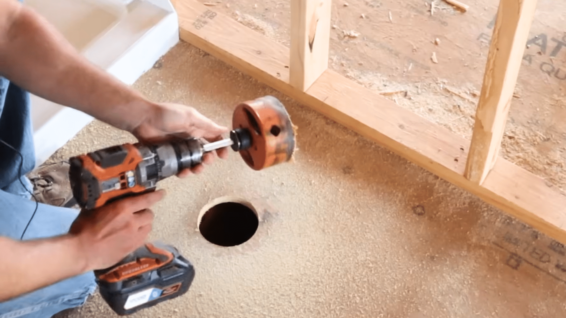hands holding power drill with hole saw attachment positioned above circular hole cut into subfloor in construction area