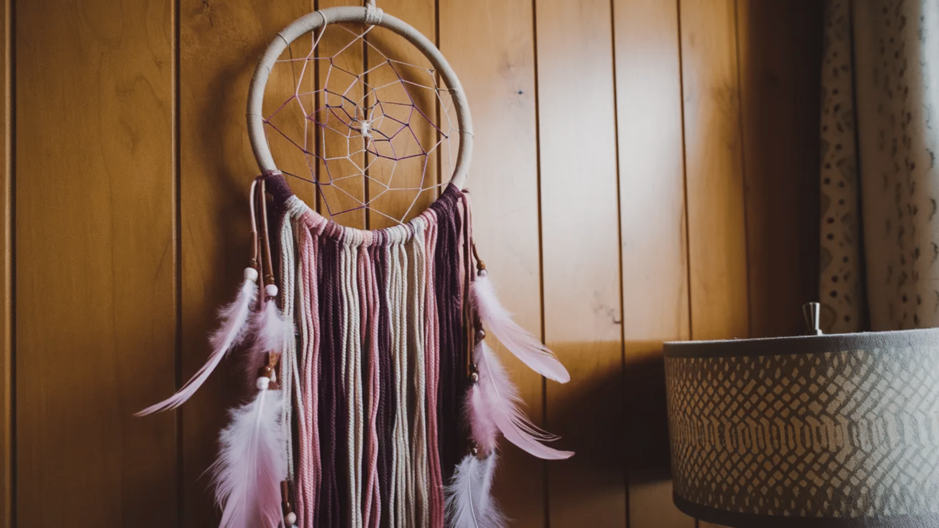 image features a beautiful dreamcatcher with soft pink, beige, and burgundy tones. The dreamcatcher hangs against a warm wooden wall, with delicate feathers and beads
