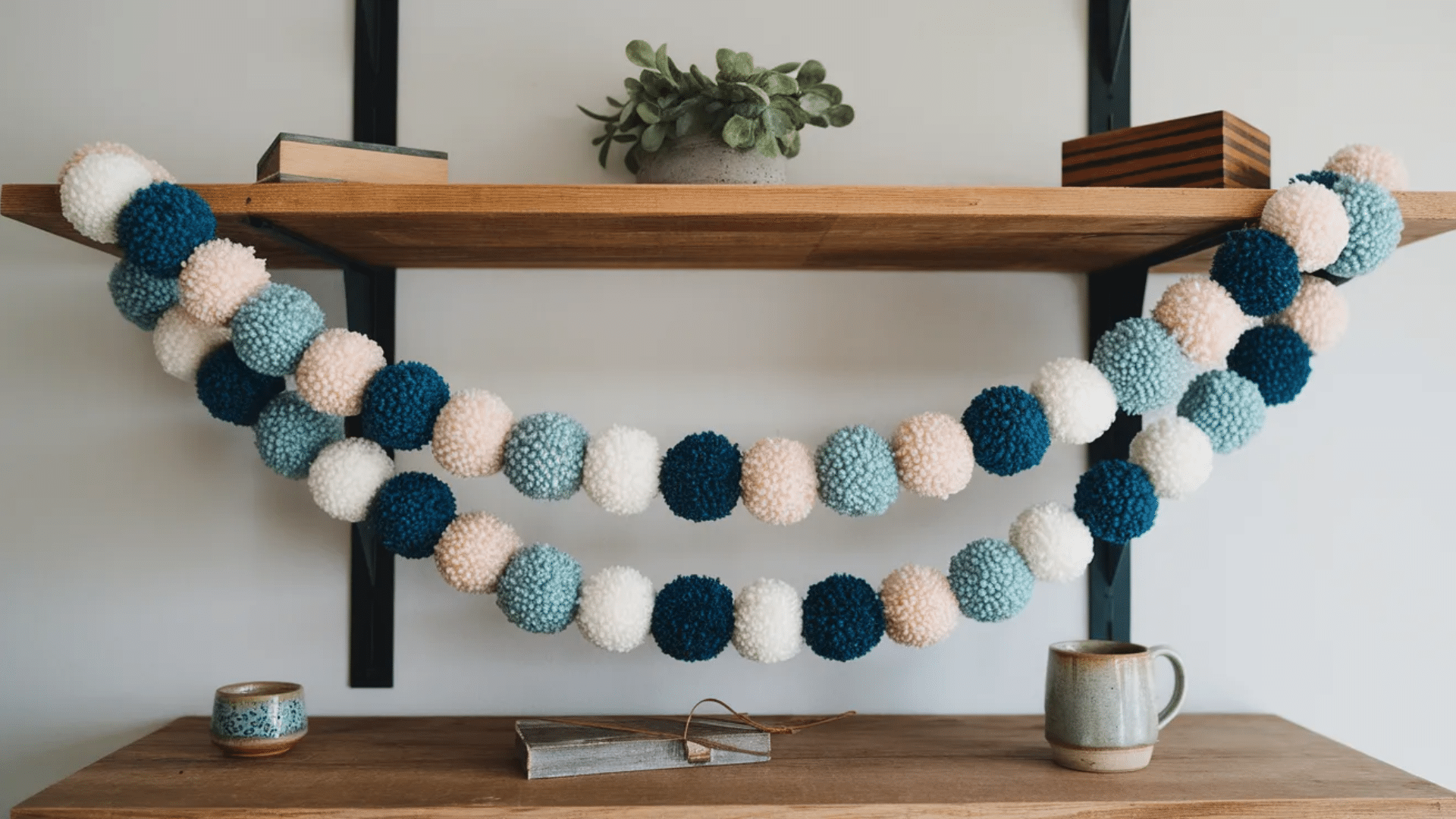 image features a shelf with a cozy and charming display. A string of colorful pom-pom garland in shades of teal, navy blue, pale pink, and white drapes across the shelf.