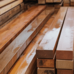 image shows neatly stacked wooden planks in a warehouse, showcasing various shades of wood, some darker and others lighter, possibly for woodworking or construction projects
