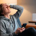 Person in gray hoodie frustrated with phone on couch in softly lit living room