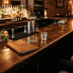long wooden bar counter with stools, drinks setup, and dim ambient lighting.