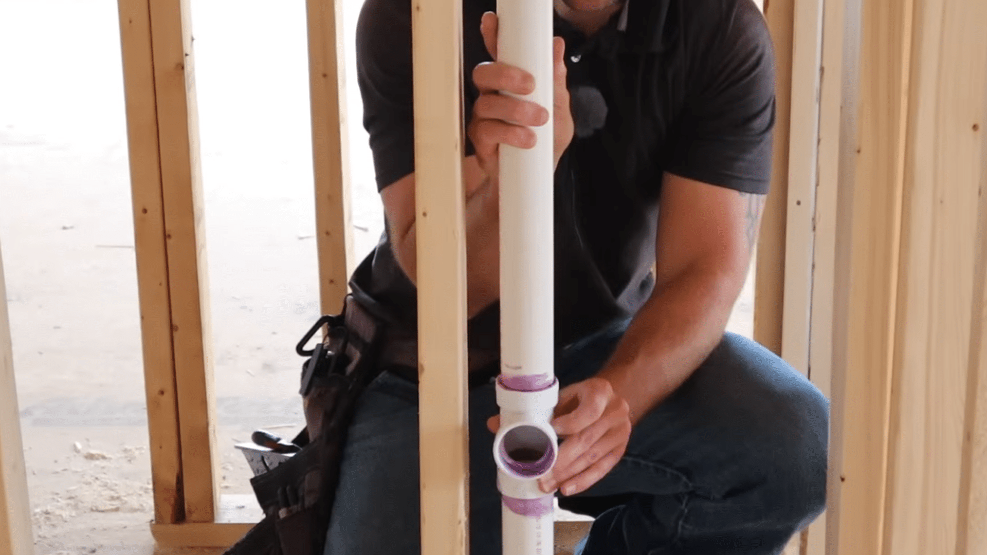 man aligning and pressing vertical pvc pipe into primed fitting inside wooden wall studs during bathroom plumbing installation