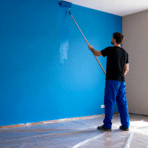 man painting interior wall with roller on extension pole applying bright blue paint in room with covered floor and window