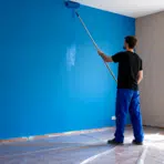 man painting interior wall with roller on extension pole applying bright blue paint in room with covered floor and window