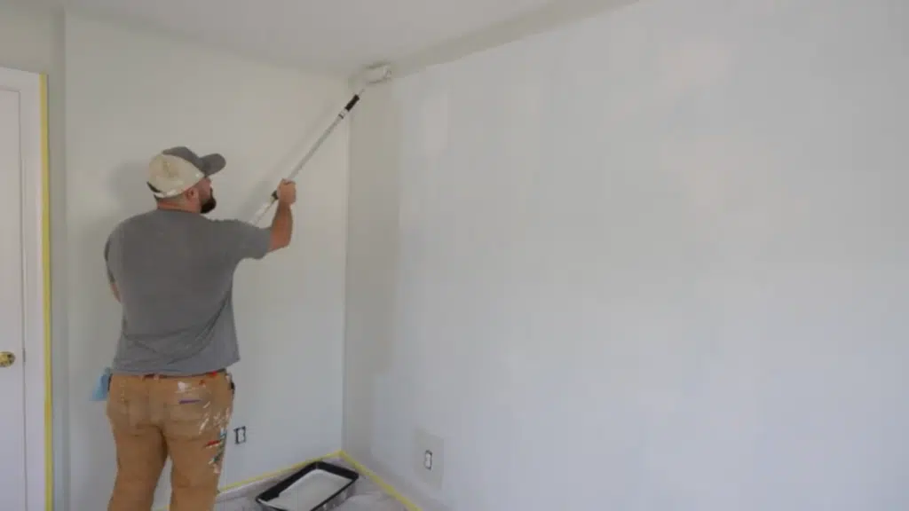 man using paint roller to apply white paint on wall in room with drop cloth and tray