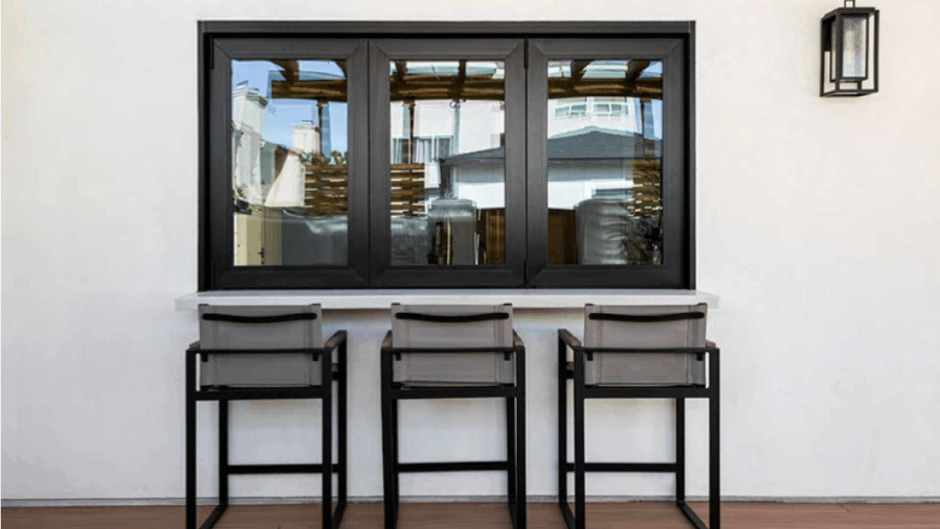 outdoor bar counter with three stools beneath black framed window, white wall backdrop, and modern patio seating setup