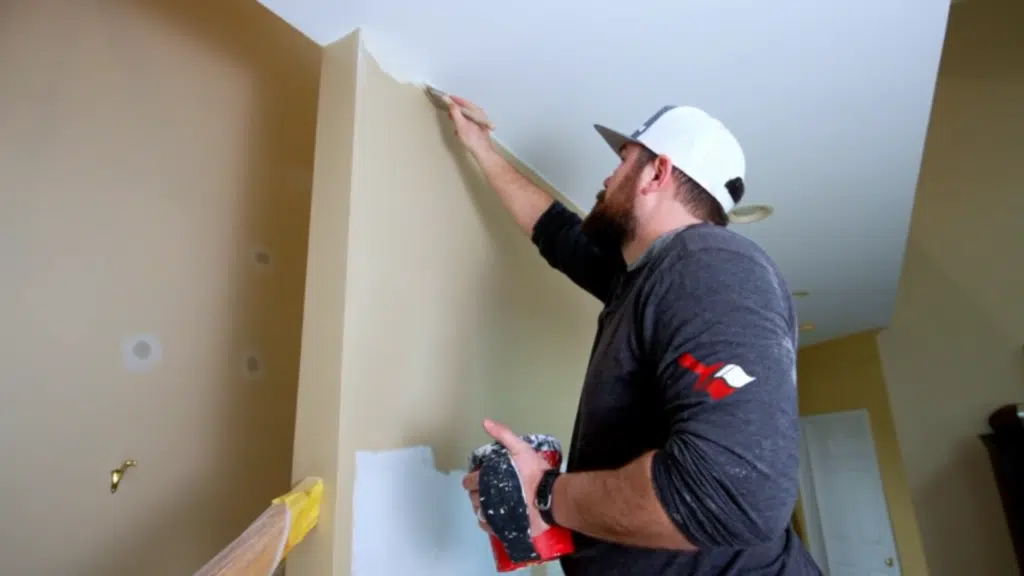 person applying paint along ceiling corner with brush on beige wall holding paint container