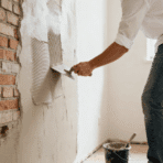 person applying plaster on a brick wall using a trowel, smoothing surface indoors with tools and bucket nearby in renovation setup