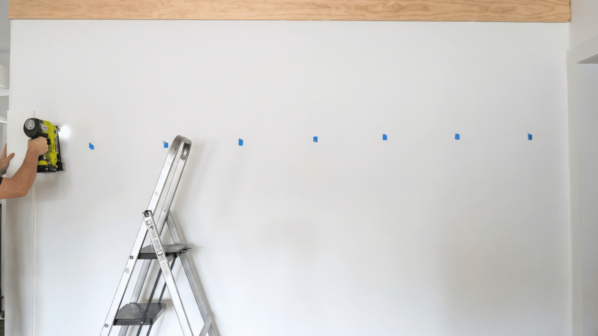 person drilling into a wall marked with blue painter's tape while standing on a ladder