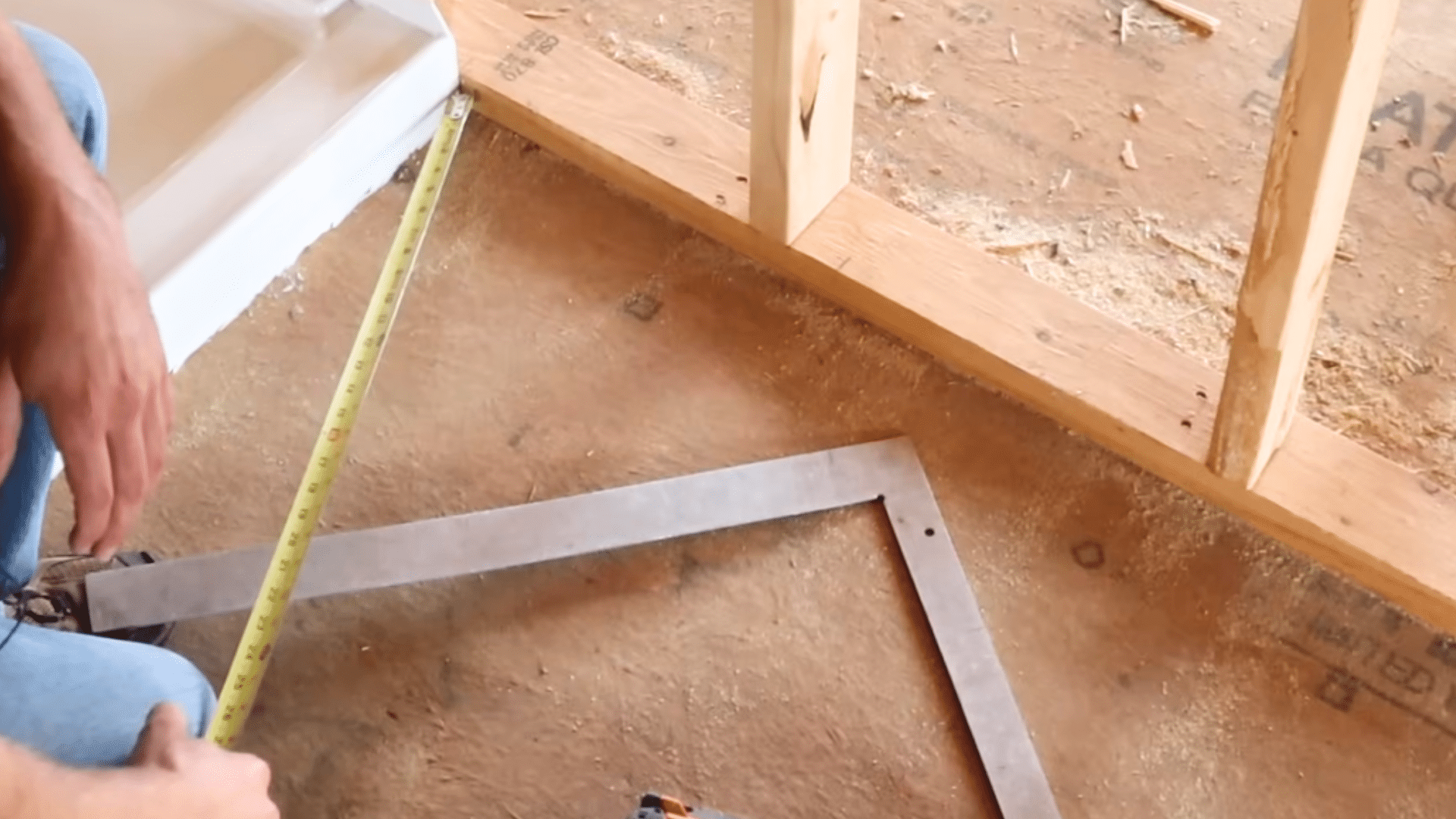 person measuring floor near wooden studs with tape measure and metal square inside unfinished room with shower base nearby