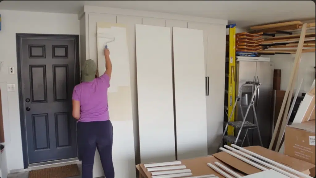 person painting vertical wall panel beside white wardrobe using roller, applying first coat while tools and materials sit nearby