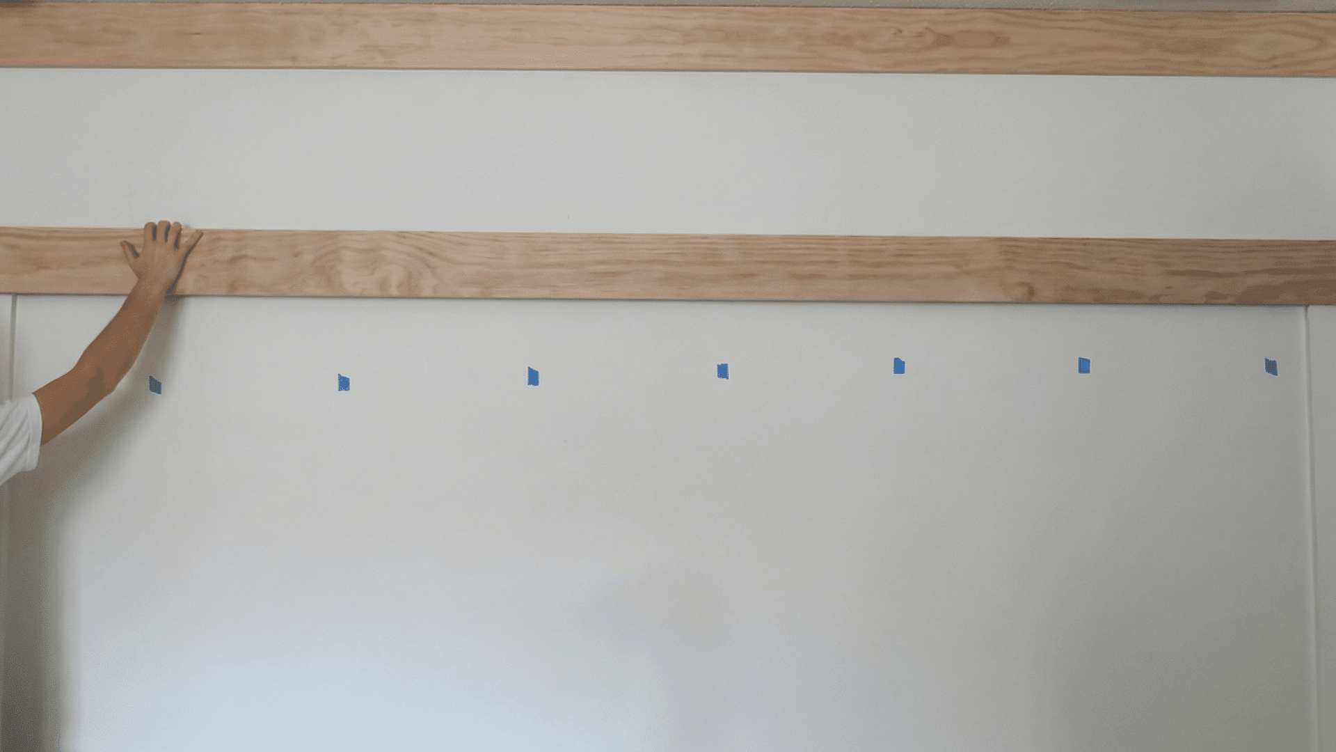 person placing a wooden board on a wall with blue painter's tape marking spots