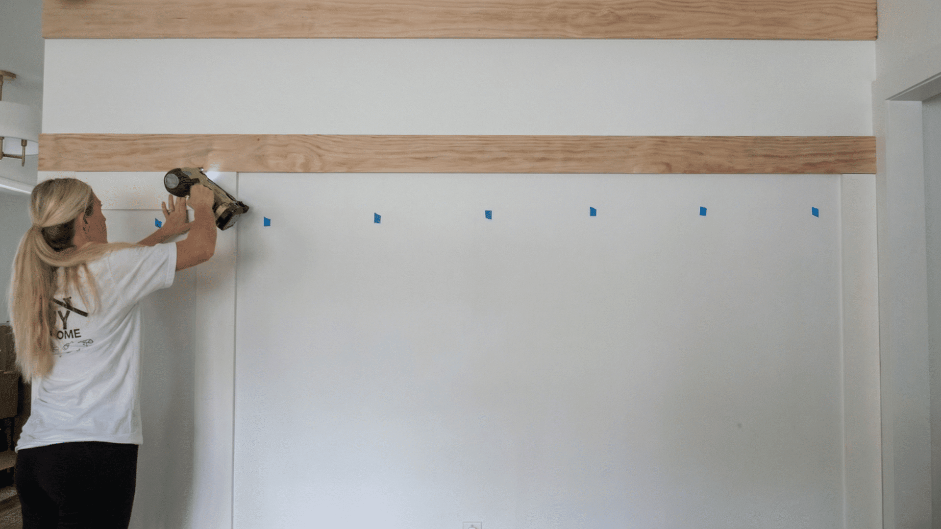 person using a nail gun to attach a wooden board to the wall marked with blue painter's tape