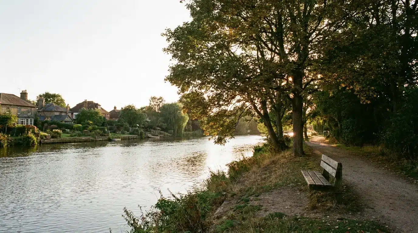 Riverside path with wooden bench under shade of large trees, village houses in background