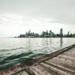 City skyline across a calm lake with wooden pier in foreground under cloudy sky