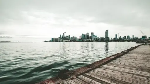 City skyline across a calm lake with wooden pier in foreground under cloudy sky