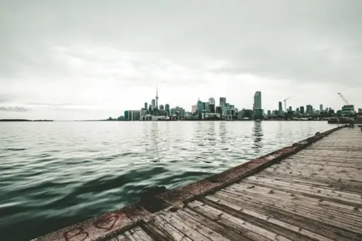 City skyline across a calm lake with wooden pier in foreground under cloudy sky