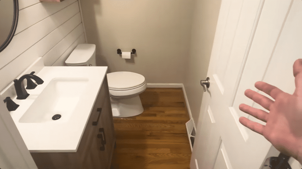 the image shows a bathroom with a white sink and black faucet, a white toilet, a toilet paper holder, and light wood flooring. The door is partially open with a hand visible holding