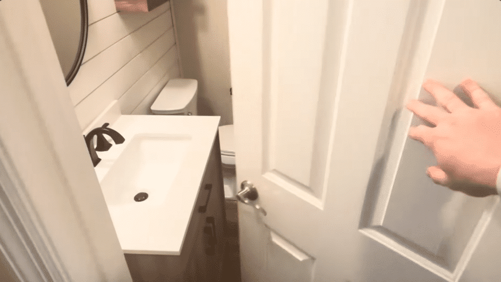 the image shows a partially open bathroom door with a hand gripping the door handle. The bathroom features a white sink, a black faucet, and a toilet in the background
