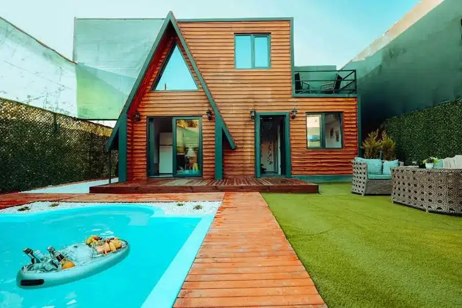 Modern wooden cabin with A-frame design, poolside seating, and artificial turf yard