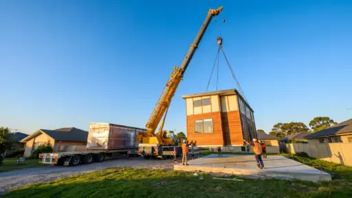Crane lifting wooden modular home onto foundation in residential area under blue sky