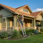 Man on ladder inspecting house gutter in sunlit suburban yard