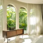 Mid-century console table by arched windows with sheer curtains, overlooking lush garden in bright room
