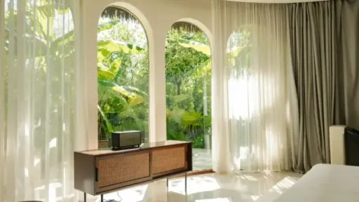 Mid-century console table by arched windows with sheer curtains, overlooking lush garden in bright room
