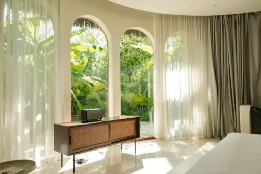 Mid-century console table by arched windows with sheer curtains, overlooking lush garden in bright room