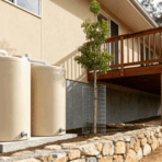 two large, beige water tanks are situated next to the house, connected by white pipes to the gutter system. A small tree, enclosed by a wire cage