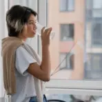 Person drinking water from glass by window with city view in bright indoor setting