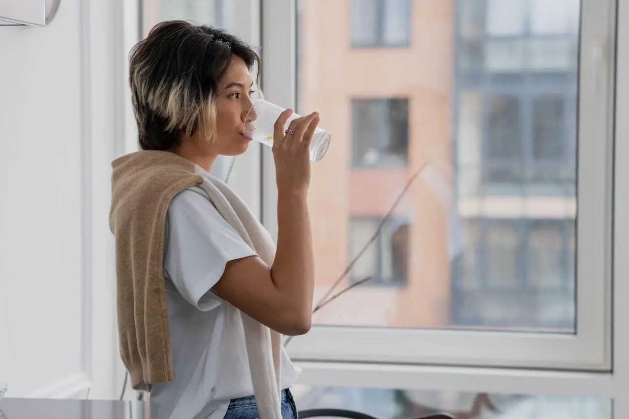 Person drinking water from glass by window with city view in bright indoor setting