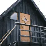 Black wooden cabin exterior with person climbing snow-dusted staircase and geometric door design