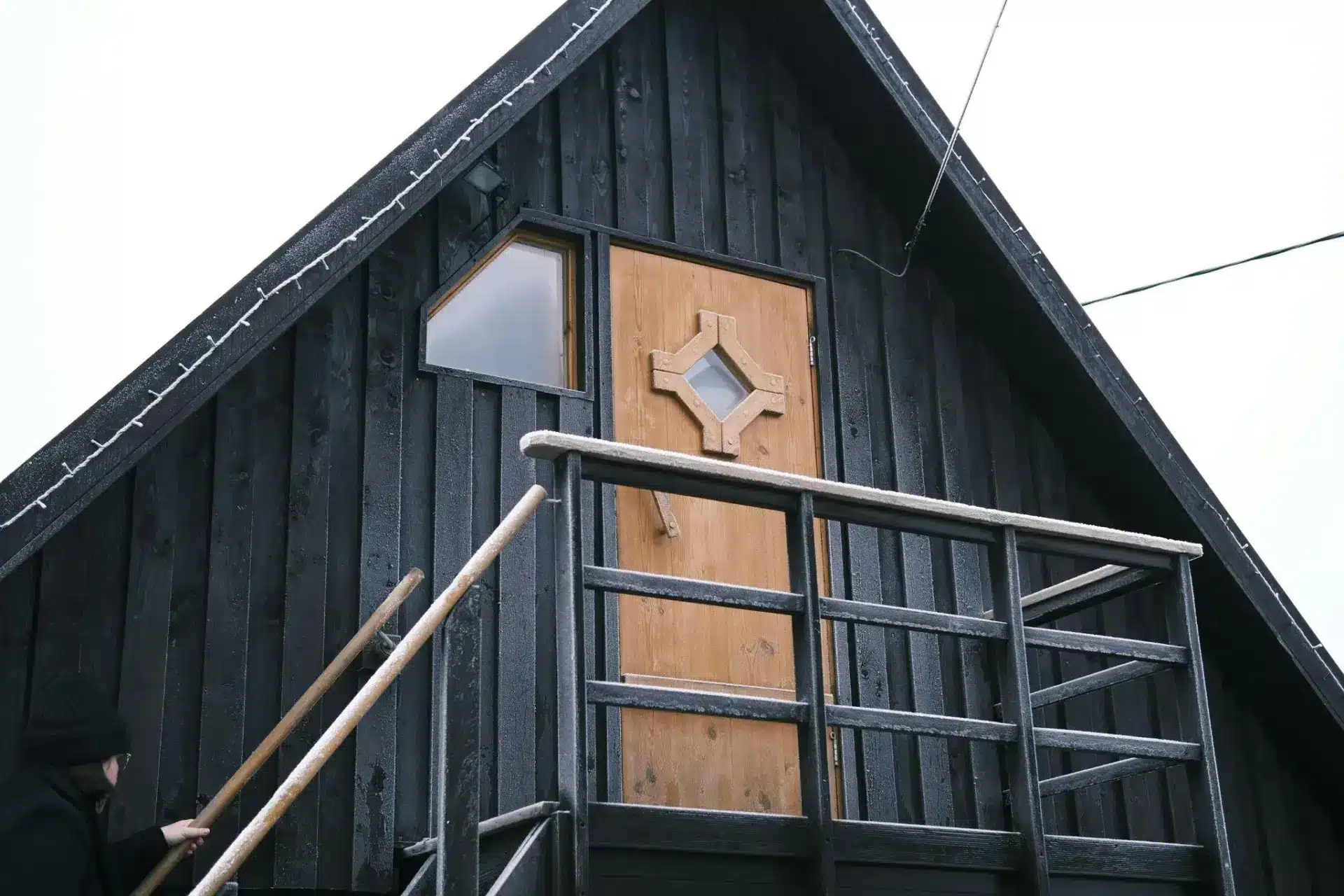 Black wooden cabin exterior with person climbing snow-dusted staircase and geometric door design